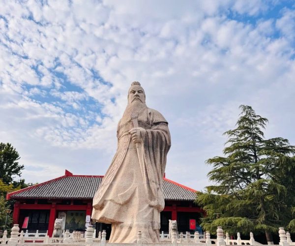 Cao Clan Cemetery,Bozhou,China tour Large stone statue of a bearded ancient scholar in flowing robes, holding a staff, in a temple courtyard with a red-roofed building behind.