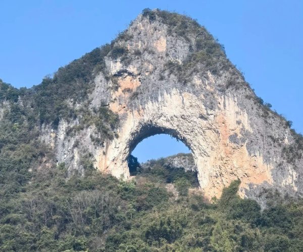 Moon Hill,Yangshuo,Chinatravel Stone mountain with a large natural arch carved through its center, set against a clear blue sky and forested lower slopes.