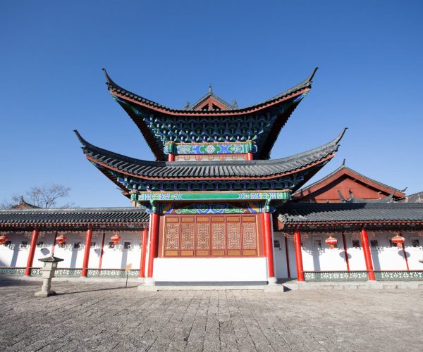 Mu’s Residence,Lijiang,Chinatravel Traditional Chinese temple gate with layered, upturned eaves, vibrant blue and green ornamentation, red pillars, and surrounding white walls under a clear blue sky
