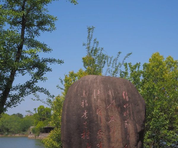 Xinghua Village,Chizhou,China tour Dark boulder with vertical red Chinese inscriptions set in a sunny park by a lake, green trees and a low stone wall in the background.