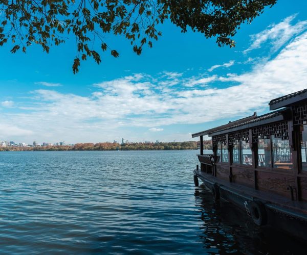 Hangzhou,Zhejiang,Chinatravel Lakeside scene with a traditional wooden pavilion along the water, under tree branches and a blue sky with distant city skyline.