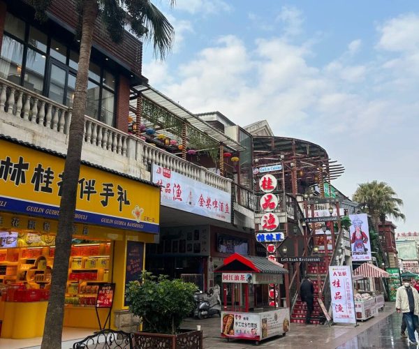 Zhengyang Pedestrian Street,Guilin,Chinatravel Busy shopping street with Chinese storefronts, a yellow shop front on the left, palm trees, and outdoor stalls under a blue sky.