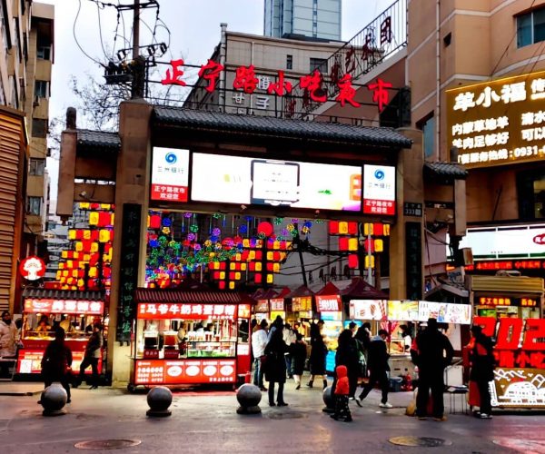 Entrance gate to a busy street market with colorful lanterns, bright signs, and food stalls; people browse and shop along the street.