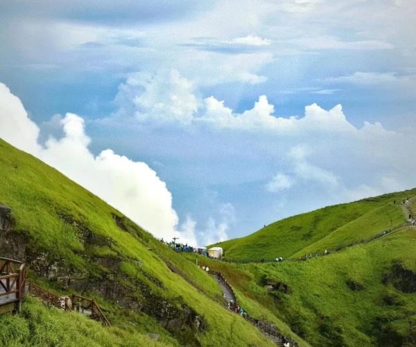 Wugong Mountain,Ji'an,China tour Winding wooden path climbs a green hillside beneath a blue sky with fluffy white clouds, hikers visible along the trail.