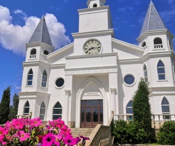 Shuishi Forest Hot Spring Resort,Qiqihar,Chinatravel White church with three spire-topped towers and a clock, under a blue sky, with pink flowers in the foreground.