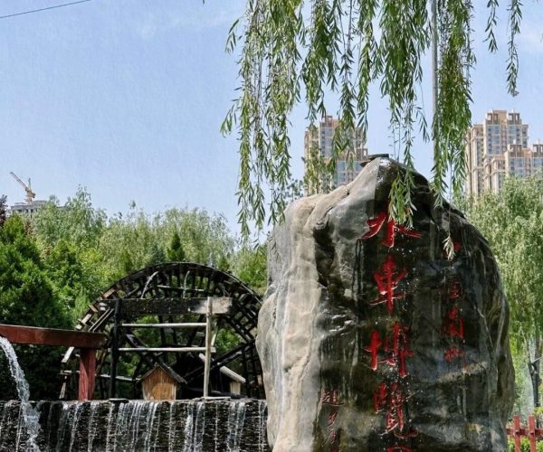 Yellow River Scenic Area & Ancient Waterwheel Park,Lanzhou,China tour Large rock with red Chinese inscriptions beside a cascading pond, willow branches overhead, and a wooden waterwheel in the background of a park.