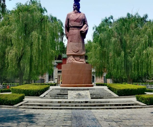 Youli Castle & Scenic Area,Anyang,Chinatravel Statue of a historical figure in traditional robes on a stone pedestal in a park with greenery and weeping trees nearby.