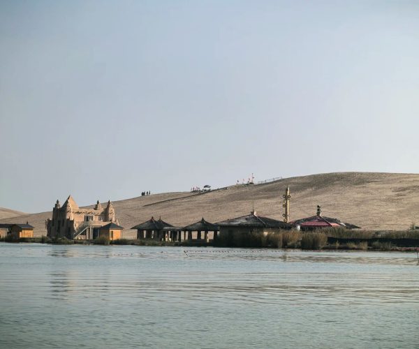 Shahu Lake (Sand Lake) Scenic Area,Yinchuan,Chinatrip Desert lakeside with a castle-like building and thatched-roof pavilions along calm water, and a sandy dune in the background.