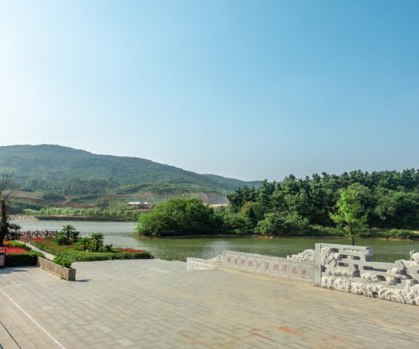 Henan Weather,Henan,Chinatravel Sunlit park featuring a calm lake, manicured shrubs, and a decorative stone railing with Chinese characters overlooking hills in the distance