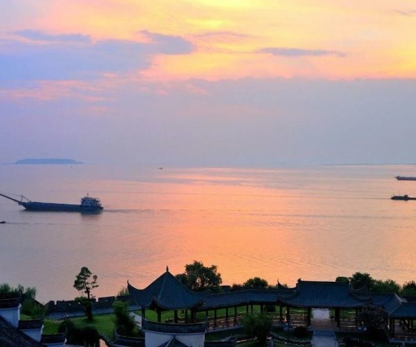 Dongting Lake,Yueyang,Chinatravel Sunset over a calm sea with pastel orange and pink sky, boats on the water, and traditional buildings in the foreground.