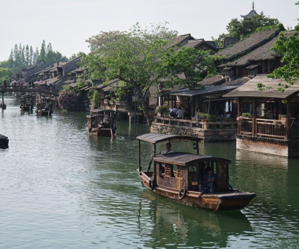 Zhejiang Weather ,Zhejiang,Chinatravel Boats travel along a canal beside traditional wooden houses and a stone bridge with people walking above and along the water's edge, surrounded by trees and plants.