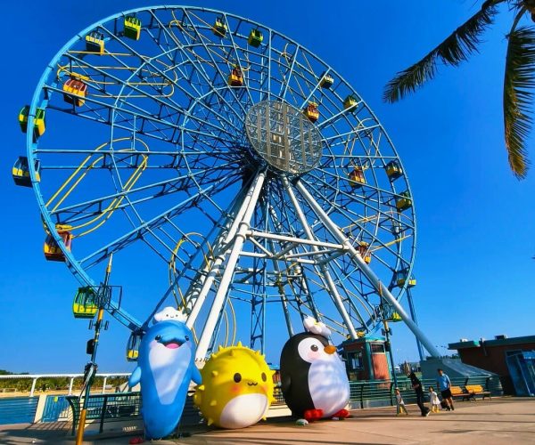 Haikou Happy World,Haikou,Chinatravel Ferris wheel at a sunny amusement park with colorful cabins and three large cartoon mascots (blue penguin, yellow sun, black penguin) at the base, palm trees in the background.