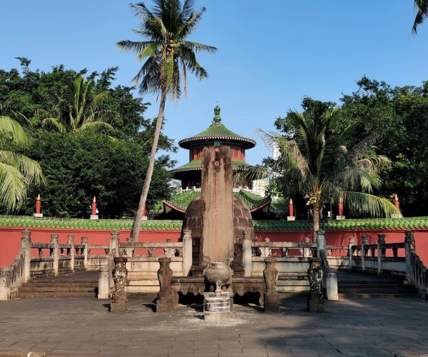 Hairui Tomb,Haikou,Chinatravel Stone pedestal monument in a courtyard with a circular pagoda-style pavilion behind, red wall with green tiled roof, and palm trees under a blue sky (outdoor temple scene)