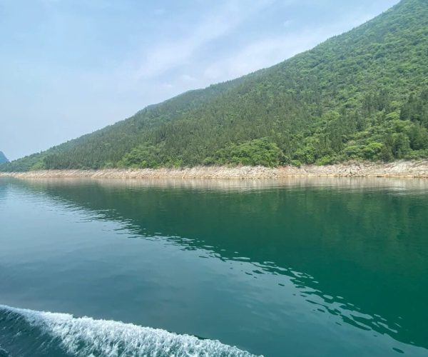 Qing River Gallery Scenic Area,Yichang,Chinatravel Calm green water near a rocky shoreline with a forested hill and a clear blue sky; boat wake visible in the foreground.