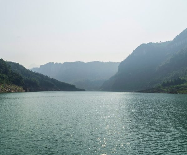 Hubei Weather,Hubei,Chinatravel Calm mountain lake with forested cliffs and hazy blue hills in the distance, sunlight glittering on the water surface.