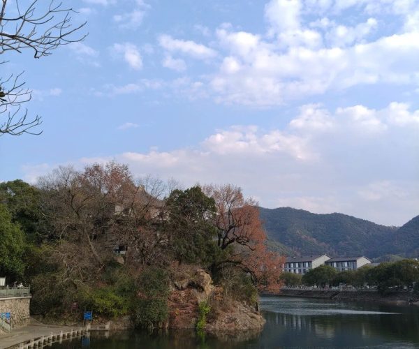 Fenghua Ancient Town,Ningbo,Chinatravel Lakeside scene with a rocky outcrop and bare trees, a stone walkway along the water, and distant buildings with hills under a partly cloudy blue sky.