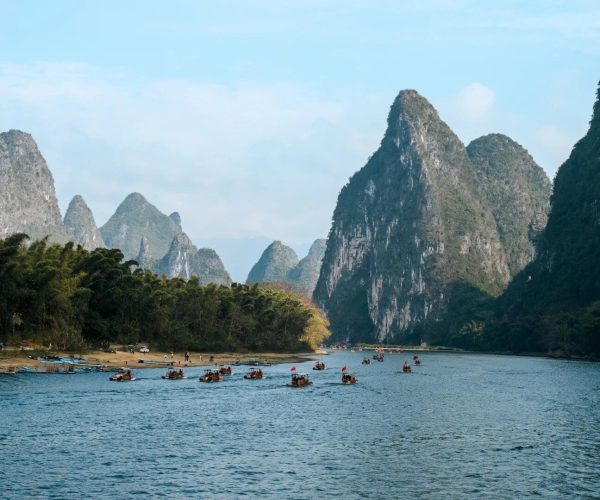 Li River,Guilin,Chinatravel Calm river lined with forested shore and groups of small boats, set against towering limestone mountains under a blue sky.