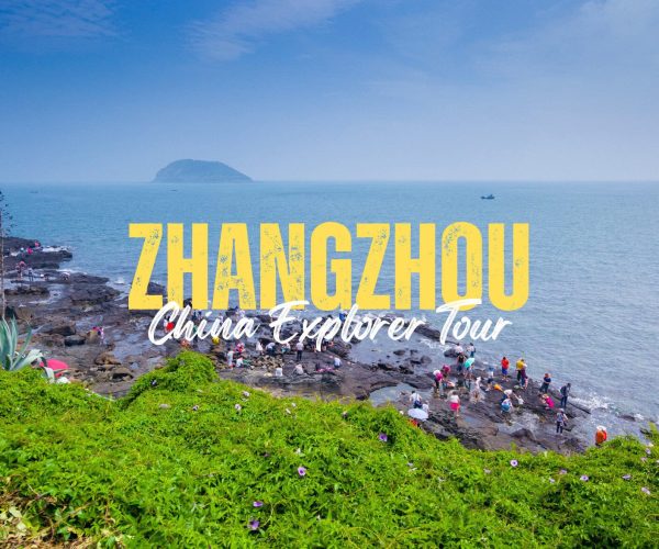 Zhangzhou,Fujian,Chinatravel Coastal rocky shore with groups of people exploring; island in the distance, blue sea and sky, green foreground, with bold yellow 'ZHANGZHOU' and white 'China Explorer Tour' text overlay.
