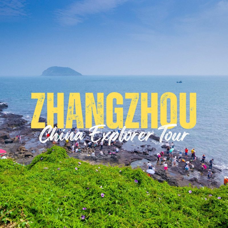 Zhangzhou,Fujian,Chinatravel Coastal rocky shore with groups of people exploring; island in the distance, blue sea and sky, green foreground, with bold yellow 'ZHANGZHOU' and white 'China Explorer Tour' text overlay.