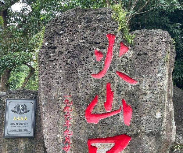Volcano Crater Park,Haikou,Chinatravel Large weathered rock with bright red Chinese characters carved into it, set among greenery and small plants along the top edge; a plaque on the left reads 'AAAA National Tourist Attraction'.