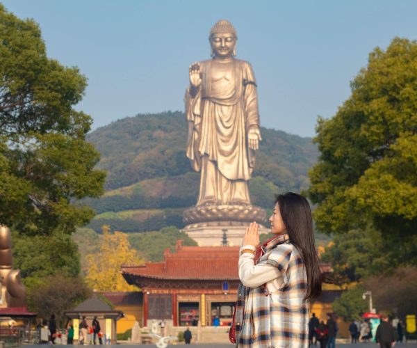 Lingshan Grand Buddha,Suzhou,Chinatravel Young woman in a plaid coat praying with pressed palms in front of a towering Buddha statue in a temple courtyard, trees in the background.