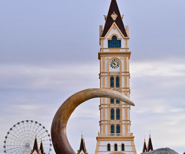 Mammoth Park,Manzhouli,China tour Tall cream clock tower with dark brown steep roof, blue windows, and decorative arches; a large curved tusk sculpture sits in the foreground, with a rock bearing Chinese characters and a Ferris wheel in the background.