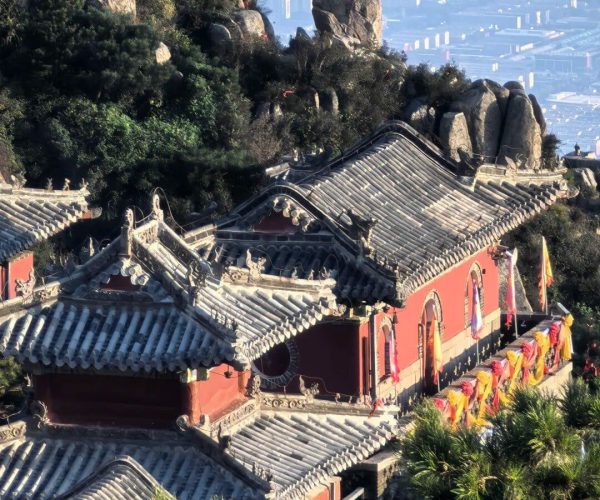Jade Emperor Peak,Tai'an,China tour Temple complex with curved gray-tiled roofs and red walls perched on a hillside, colorful banners lining the façade, against rocky cliffs and a distant city skyline.