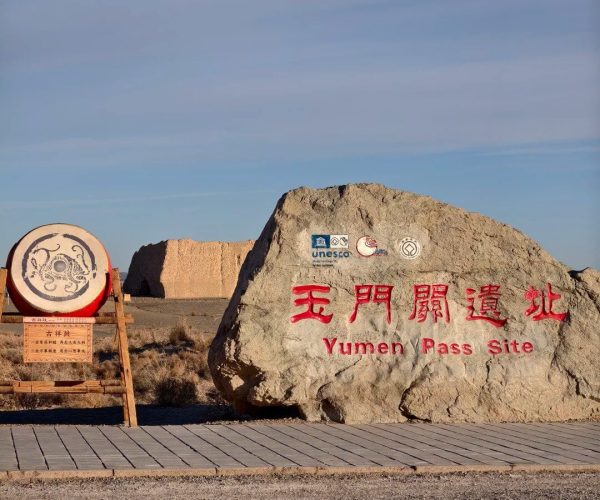 Yumen Pass,Dunhuang,Chinatrip Large rock with red Chinese characters and the words 'Yumen Pass Site' in English, with logos above, beside a wooden drum in a desert landscape.