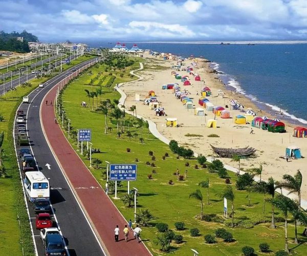 Island Ring Road,Xiamen,Chinatravel Coastal park beside a curved road with cars, green lawns and palm trees, and a busy sandy beach with colorful tents along the shoreline.