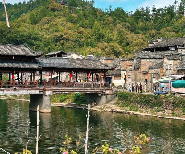 Yaoli Ancient Town,Jingdezhen,China tour Wooden covered bridge over a calm canal in a historic village, with stone buildings and a forested hill backdrop.