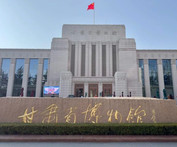 Gansu Provincial Museum,Lanzhou,China tour Front view of a large museum with a red Chinese flag atop, light stone facade and tall vertical windows; a stone sign with gold Chinese characters in the foreground.