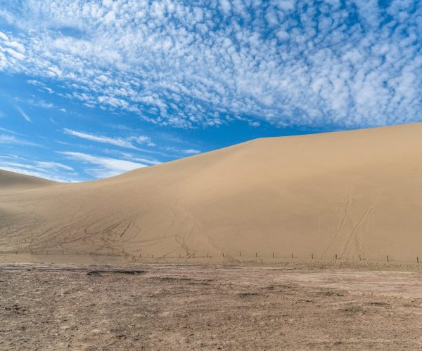 Gansu Weather,Gansu,China tour Golden desert scene with a large sand dune under a bright blue sky and scattered clouds; a fence runs along the dry, dusty foreground.