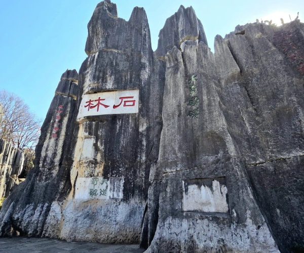 Stone Forest,Kunming,Chinatravel Jagged gray rock wall with red and green Chinese characters painted on signs, a stone-paved ground in the foreground and a clear blue sky above.