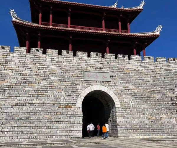 Shishi Ancient Town,Quanzhou,Chinatravel Stone fortress gate with an arched entrance and a red, multi-story pavilion atop, set against a blue sky.