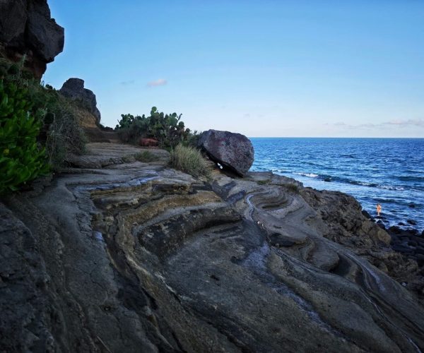 Naozhou Island,Zhanjiang,Chinatravel Rocky coastal path with layered rocks, green shrubs, and the blue ocean to the right; a person stands near the water’s edge.