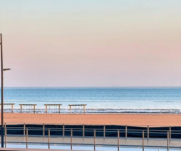 Qinhuangdao Weather,Qinhuangdao,Chinatravel Serene beach scene with a pastel sky, calm blue ocean, and empty benches along the shore behind a railing.