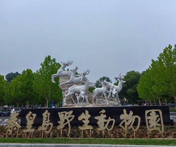 Qinhuangdao Safari Park,Qinhuangdao,Chinatravel Group of white deer statues arranged on a rock pedestal at the entrance of Qinhuangdao Wild Animal Park, in front of a large black sign with golden Chinese characters of the park’s name.
