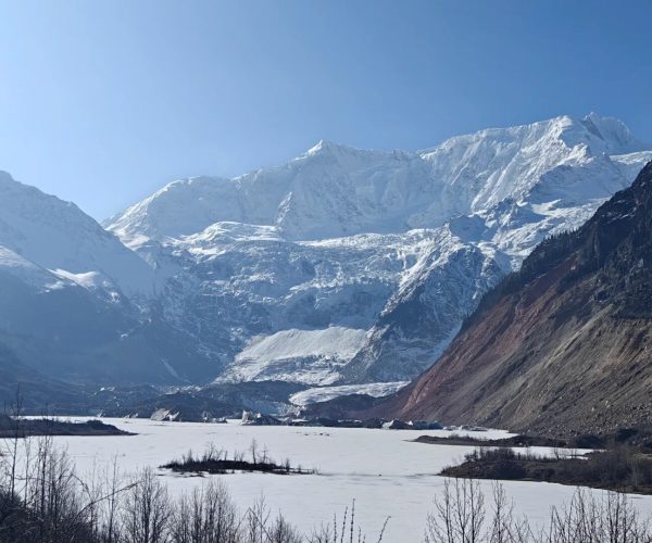 Midui Glacier,Nyingchi,China tour Snowy mountain range under a clear blue sky, with a frozen lake in the valley and bare shrubs in the foreground.