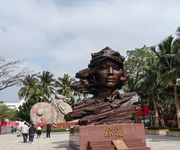 Red Detachment of Women Memorial,Qionghai,Chinatravel Bronze bust of a woman on a red granite pedestal with gold Chinese characters, set in a plaza with palm trees and a circular stone sculpture featuring red stars in the background.