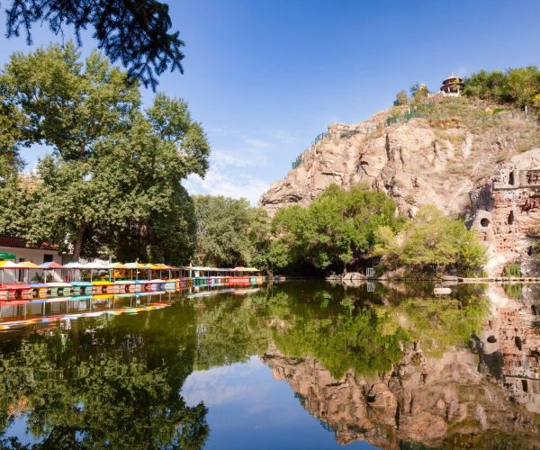 Red Hill Park,Urumqi,Chinatrip Row of colorful paddle boats docked along a tree-lined lakeshore with a rocky hill and pavilion above, reflecting in calm water.