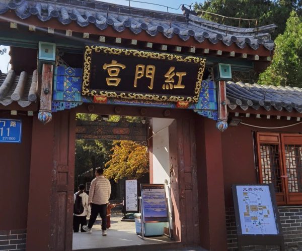 Red Gate Palace Scenic Area,Tai'an,China tour Traditional Chinese temple gate with a gold-on-black plaque, colorful decorative trim, and a stone-paved entry leading to a courtyard behind.Two people and a child walk through the gateway on a sunny day.