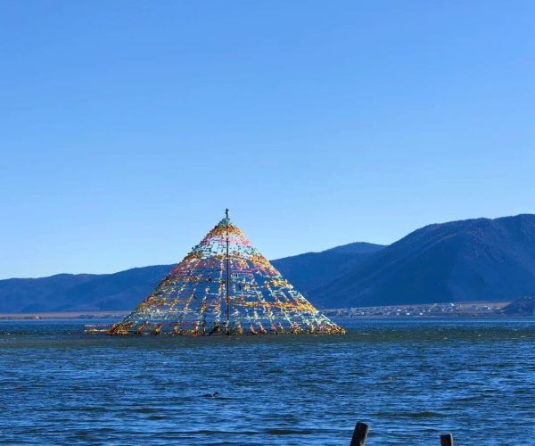 Napahai Lake,Shangri-La,Chinatravel Colorful pyramid-shaped structure made of hanging prayer flags rising from a calm lake with distant mountains under a clear blue sky.