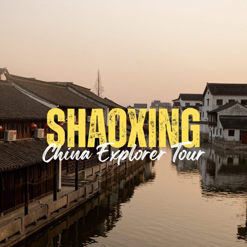 Shaoxing,Zhejiang,Chinatravel Promotional banner for a Shaoxing China Explorer Tour with bold yellow 'SHAOXING' text over a canal-side town at sunset, with a cursive 'China Explorer Tour' subtitle is visible over the water.