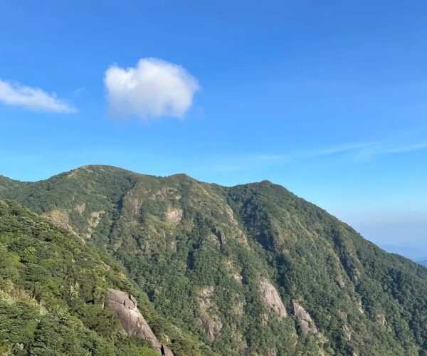 Luofu Mountain,Huizhou,Chinatravel Green, forested mountains with rocky ridges under a clear blue sky and a small white cloud above the ridge.
