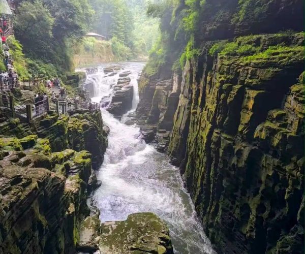 Tenglong Cave,Enshi,Chinatravel Narrow rocky gorge with a rushing waterfall and mossy cliffs, a wooden walkway along the left edge where visitors stand among green forested walls.