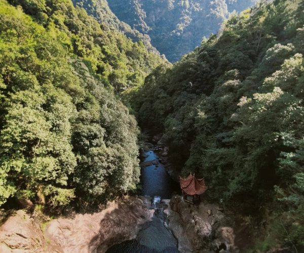 Huayan National Forest Park,Wenzhou,Chinatravel A narrow river winds through a lush green gorge with towering forested walls and a small red-roofed gazebo on the rocks below.