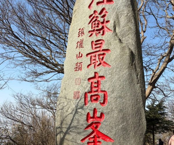 Huaguo Mountain,Lianyungang,China tour Tall gray stone monument with vertical red Chinese characters carved/painted on its front, set in a park with bare trees and a blue sky; a few people near the base on the right edge.
