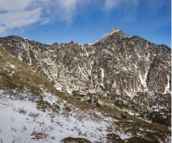 Cangshan Mountain ,Dali,Chinatravel Snow-covered mountain ridge under a clear blue sky, with sparse trees on the snowy slope in the foreground.