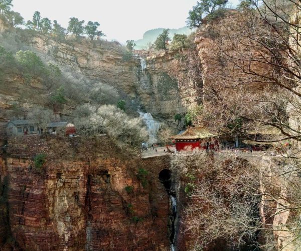 Cangyan Mountain Scenic Area,Shijiazhuang,China tour Cliffside canyon with red rock walls, a small red pavilion on a walkway, and a waterfall streaming down the cliff amid bare trees.