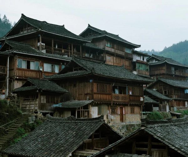Qinglong Ancient Bridge & Miao Architecture,Fenghuang,Chinatravel Traditional wooden village houses with tiled roofs line a hillside, connected by stone steps and balconies, with forested mountains in the background.